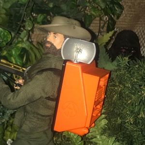 May include: A man wearing a brown hat and green jacket is carrying an orange backpack with a black strap and a silver antenna. The backpack has a black and white logo with the letter "A" inside a circle. The backpack has a black and white screen with a grid pattern.