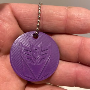 May include: A purple keychain with a 3D printed design of the Decepticons logo.