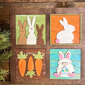 May include: A square wooden wall sign featuring four distinct panels. The top left shows three rabbits on green, the top right a white rabbit on orange with carrots, the bottom left three orange carrots, and the bottom right a gnome holding an egg.
