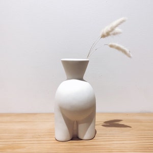 May include: White ceramic vase with a unique, human-like shape. The vase has a wide, round base and two legs. It is filled with dried white pampas grass.