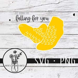 May include: A yellow graphic design of two socks with the text "At risk of falling for you" above the socks. The graphic is on a white wood background with a black and white floral design in a circle at the bottom of the image. The text "SVG - PNG" is at the bottom of the image.