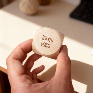 Könnte beinhalten: Ein Holzwürfel mit den Worten "ICH BIN GENUG" auf einer Seite eingraviert. Der Würfel wird in einer Hand gehalten, mit einem neutralen Hintergrund. Der Text ist in einer einfachen, serifenlosen Schriftart.