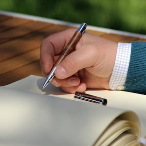 Könnte beinhalten: Eine Hand hält einen Holzstift mit der Gravur "Matthias". Der Stift wird verwendet, um in ein leeres Notizbuch zu schreiben.