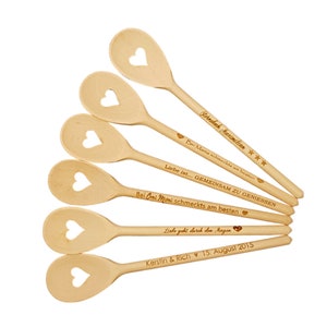 Op de afbeelding: Zes houten lepels met hartvormige uitsnijdingen. Elke lepel is gegraveerd met een gepersonaliseerd bericht, waaronder "Sternelch Maximilian", "Bei Mama schmeckts am besten", "Liebe ist... GEMEINSAM ZU GENIESSEN", "Bei Omi Moni schmeckts am besten", "Liebe geht durch den Magen" en "Kerstin & Rich 15. August 2015".