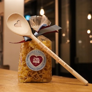 Op de afbeelding: Een houten lepel met een gegraveerd hart, vastgebonden aan een zak met hartvormige pasta. De zak heeft een sticker met de tekst "Danke f&uuml;r dein Herz" en "Zusammen Gl&uuml;cklich". De lepel is gegraveerd met de tekst "Bring die Ideenk&uuml;che zum Brodeln" en "Herz Caspar".