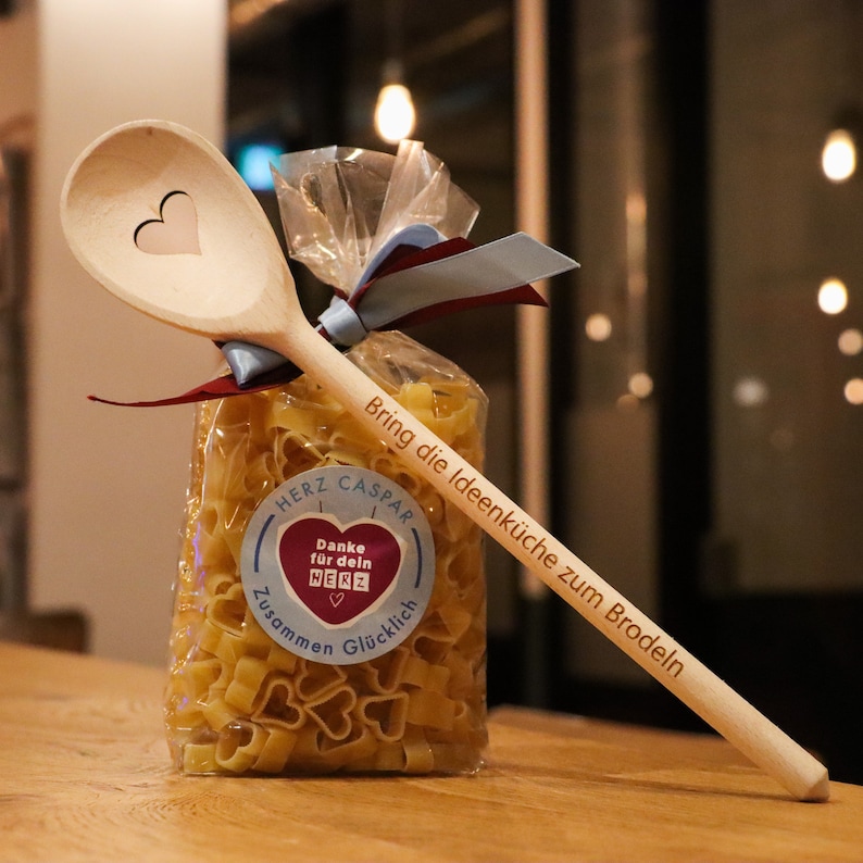 Op de afbeelding: Een houten lepel met een gegraveerd hart, vastgebonden aan een zak met hartvormige pasta. De zak heeft een sticker met de tekst "Danke f&uuml;r dein Herz" en "Zusammen Gl&uuml;cklich". De lepel is gegraveerd met de tekst "Bring die Ideenk&uuml;che zum Brodeln" en "Herz Caspar".
