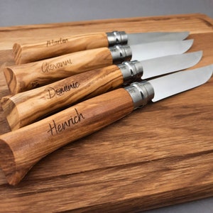 Könnte beinhalten: Vier Messer mit Holzgriffen und personalisierten Namen: Martin, Giovanni, Dominic und Heinrich. Die Messer liegen auf einem Holzbrett.