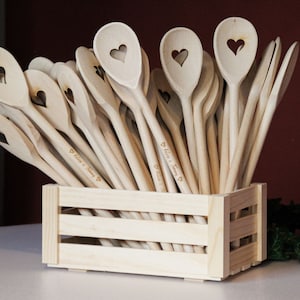 Op de afbeelding: Een houten kist gevuld met houten lepels, elk met een hartvormige uitsparing en gepersonaliseerd met de tekst "Felix & Jenny" in cursief.