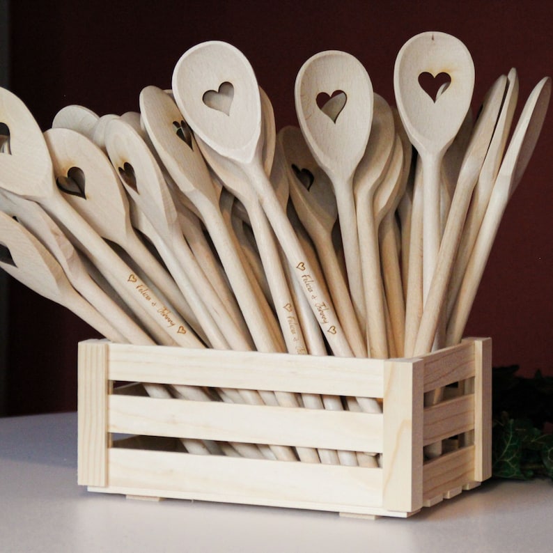 Op de afbeelding: Een houten kist gevuld met houten lepels, elk met een hartvormige uitsparing en gepersonaliseerd met de tekst "Felix & Jenny" in cursief.