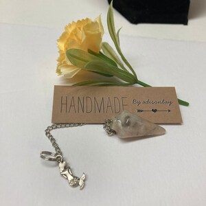Puede incluir: Un péndulo de cristal blanco y gris con una cadena plateada y un dije de sirena. El péndulo cuelga de una tarjeta marrón con el texto "HANDMADE by adisonbay".