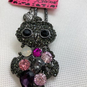 Può includere: Una collana di metallo nero a forma di gufo con una catena. Il gufo è decorato con strass di vari colori, tra cui nero, viola, rosa e trasparente. La collana ha un piccolo ciondolo a forma di cuore con le parole "Love You" su di esso. L'etichetta attaccata alla collana dice "Betsey Johnson".