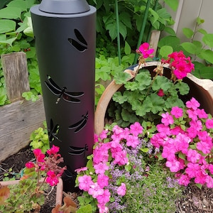 Peut inclure: Lampe de jardin solaire en métal noir avec un motif de libellule. La lumière est entourée de fleurs roses et de plantes vertes.