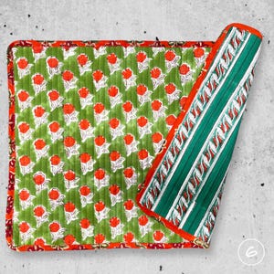 May include: A square quilted fabric mat featuring a green background with a repeating pattern of orange flowers and white leaves. The border is a dark red floral print, and one corner is folded back to reveal a green and white striped underside.