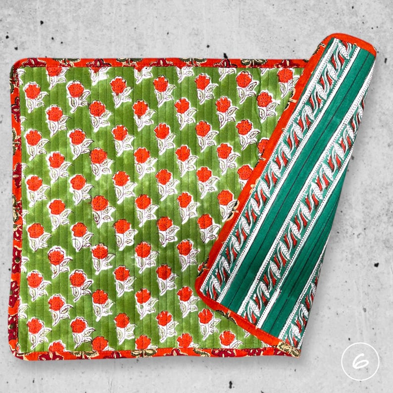 May include: A square quilted fabric mat featuring a green background with a repeating pattern of orange flowers and white leaves. The border is a dark red floral print, and one corner is folded back to reveal a green and white striped underside.