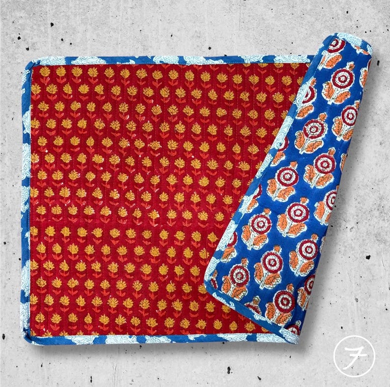 May include: A rectangular quilted fabric mat featuring a deep red base with a repeating pattern of small yellow flowers. The mat has a blue border with a white floral print, and one corner is folded back to reveal a blue underside with large floral motifs.