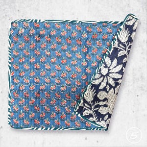 May include: A square quilted textile featuring a blue background with a repeating pattern of small red and white flowers. The corner is folded back to reveal a contrasting dark blue and white floral print with large botanical motifs.