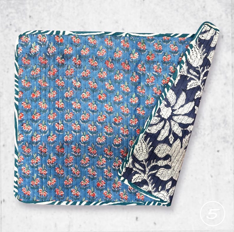 May include: A square quilted textile featuring a blue background with a repeating pattern of small red and white flowers. The corner is folded back to reveal a contrasting dark blue and white floral print with large botanical motifs.