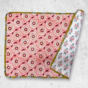 May include: A rectangular quilted blanket featuring a pink background with a repeating pattern of white and dark red circular floral motifs. The edges are finished with a mustard yellow border, and the folded corner reveals a white floral print.