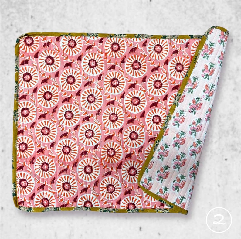 May include: A rectangular quilted blanket featuring a pink background with a repeating pattern of white and dark red circular floral motifs. The edges are finished with a mustard yellow border, and the folded corner reveals a white floral print.