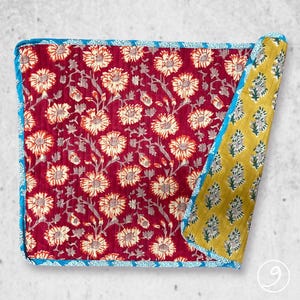 May include: A square quilted textile featuring a deep red background with a repeating cream-colored floral pattern. The piece has a bright blue patterned border and a folded corner revealing a mustard yellow underside with a small floral print.