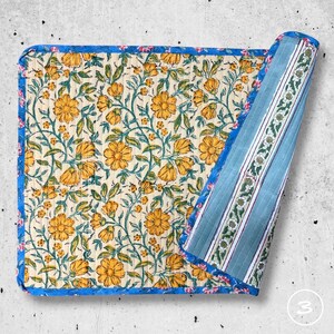 May include: A square quilted fabric mat featuring a cream background with a repeating pattern of yellow flowers and green vines. The mat has a bright blue floral border and is folded to reveal a reverse side with blue and white vertical stripes.