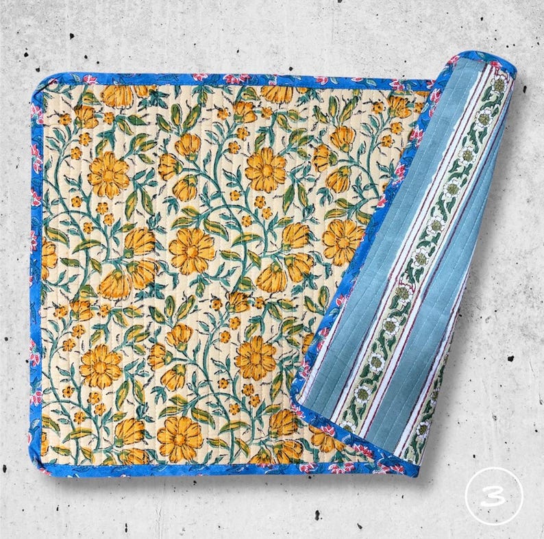 May include: A square quilted fabric mat featuring a cream background with a repeating pattern of yellow flowers and green vines. The mat has a bright blue floral border and is folded to reveal a reverse side with blue and white vertical stripes.