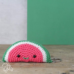 May include: A crocheted watermelon slice keyring with a smiling face. The watermelon is red with white and green stripes.