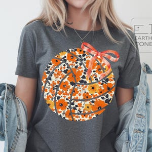 May include: A grey t-shirt with a floral volleyball design. The design features orange and yellow flowers, black detailing, and a red bow. The shirt has a crew neck and short sleeves. The Earth Tone logo is visible in the upper right corner.