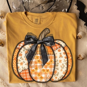 May include: Mustard-colored Comfort Colors t-shirt featuring a large pumpkin design. The pumpkin has a black bow and is decorated with floral and plaid patterns in orange, cream, and white. A black lace trim outlines the pumpkin.