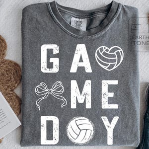 May include: Dark grey t-shirt featuring the words "GAME DAY" in distressed white lettering. The design includes a volleyball heart, a bow, and a volleyball. The shirt is made of a soft, comfortable material.