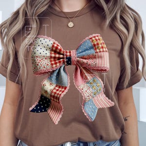 Op de afbeelding: Bruin t-shirt met een groot patchwork strik ontwerp. De strik bevat rode, blauwe en roze stoffen met bloemen- en sterrenpatronen. De strik heeft een kanten rand en is gecentreerd op het t-shirt.