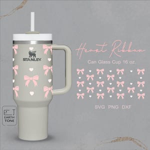 Op de afbeelding: Een lichtgrijze Stanley-beker met een roze hart- en strikpatroon. De beker heeft een handvat en een rietje. De tekst "Heart Ribbon" en "Can Glass Cup 16 oz." staat op de afbeelding. De tekst "SVG PNG DXF" staat ook op de afbeelding.