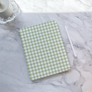 Puede incluir: Una funda para tableta con estampado de cuadros vichy verde y blanco y un lápiz óptico blanco sobre una superficie de mármol. La funda de la tableta es rectangular y tiene un diseño de cuadros clásico. El lápiz óptico es largo y delgado, colocado junto a la funda.