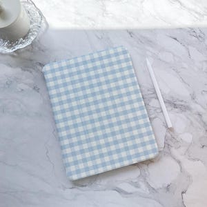 May include: A light blue and white gingham pattern tablet case with a white stylus. The case is rectangular with rounded corners. The background is a marble-effect surface.