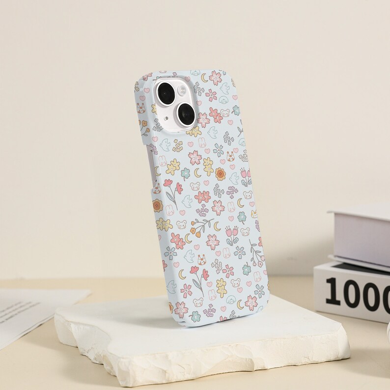 May include: A light blue phone case with a cute pattern of flowers, animals, and moons. The case is made of a soft, flexible material and has a raised lip around the camera lens to protect it from scratches.