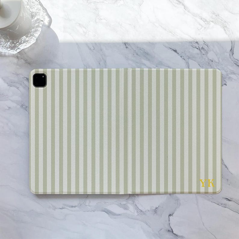 May include: A light green and white striped tablet case with a camera cutout. The case has the initials "YK" in gold on the bottom right corner. The case is rectangular and has a smooth surface.