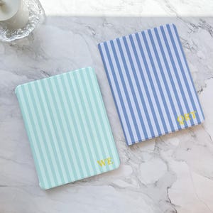 May include: Two rectangular cases with vertical stripes. One is light blue and white, the other is blue and white. Each case has gold lettering in the corner. The cases are on a marble surface.