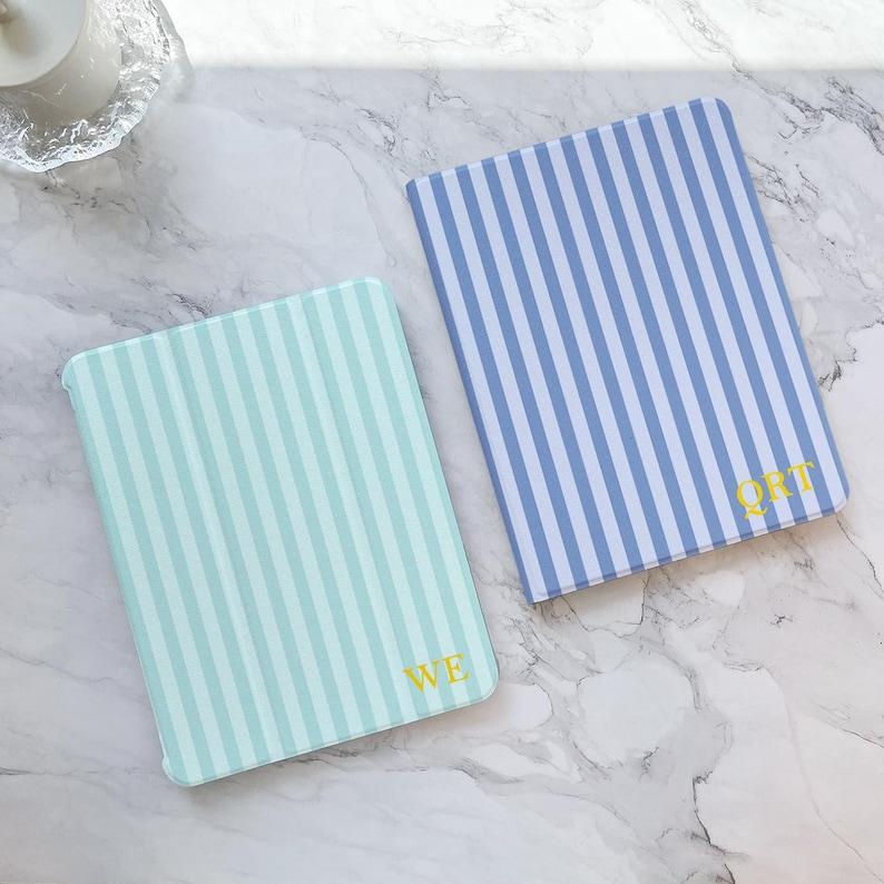 May include: Two rectangular cases with vertical stripes. One is light blue and white, the other is blue and white. Each case has gold lettering in the corner. The cases are on a marble surface.