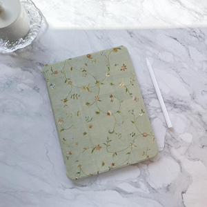 Puede incluir: Una funda de tableta verde claro con un delicado diseño floral con pequeñas flores rosas y naranjas y vides verdes. Un lápiz óptico blanco descansa sobre una superficie de mármol. La funda es rectangular con esquinas redondeadas.