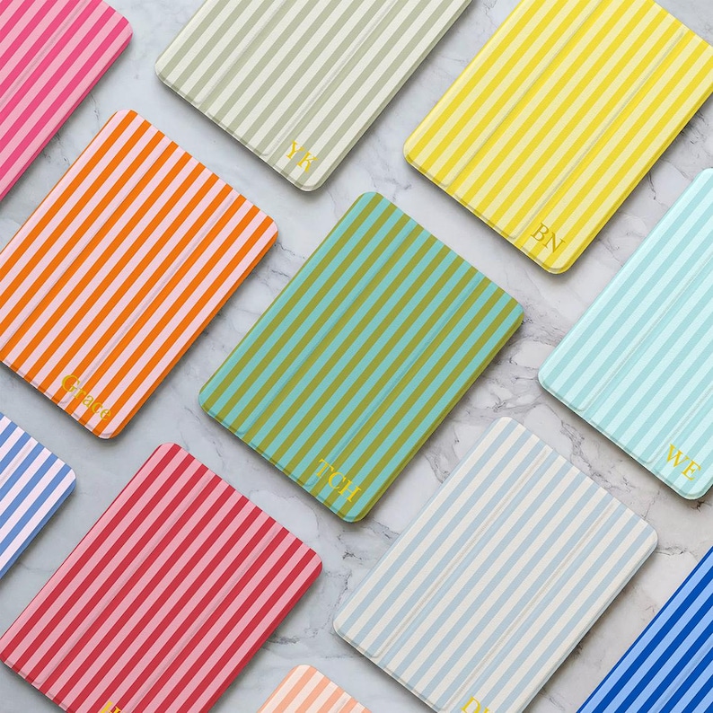 May include: A collection of rectangular cases with vertical stripes in a variety of colours, such as pink, orange, yellow, green, blue, and grey. Some cases feature gold initials. The cases are arranged on a marble surface.