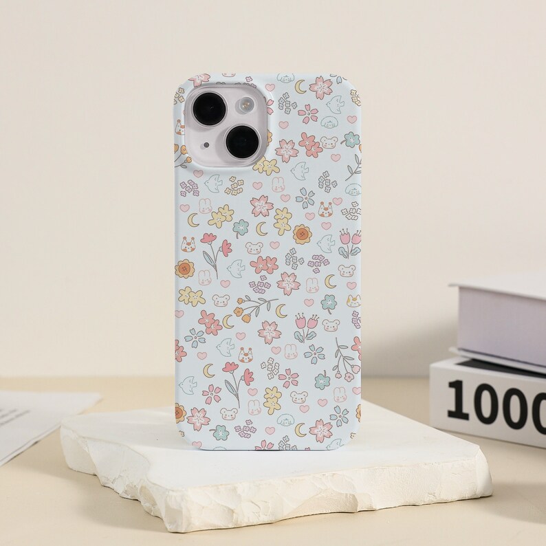 May include: A white phone case with a cute floral pattern featuring small animals, hearts, and moons. The case has a camera cutout and a light blue background.
