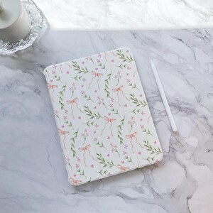 May include: A white iPad case with a floral pattern featuring pink bows, green leaves, and small pink hearts.