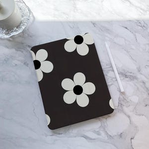 Puede incluir: Una funda negra para tableta con un patrón repetido de grandes flores blancas con centros negros. Un lápiz óptico blanco descansa sobre una superficie de mármol junto a la funda. La funda es rectangular y está diseñada para proteger una tableta.