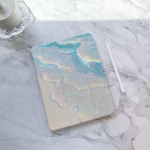 Op de afbeelding: Een tablet hoesje met een golfpatroon in blauw, wit en beige. Een witte stylus ligt naast het hoesje op een marmeren oppervlak. Het hoesje heeft een gladde afwerking en een strandachtige uitstraling. Afmetingen: ongeveer 20 cm x 15 cm.