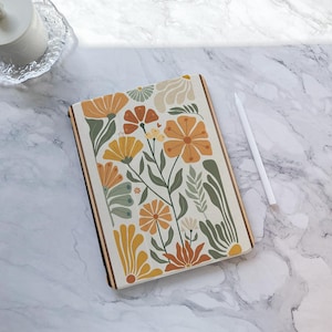 Può includere: Una custodia rettangolare per tablet con un motivo floreale nei toni dell'arancione, giallo e verde su sfondo crema. La custodia è appoggiata su una superficie di marmo accanto a uno stilo bianco.