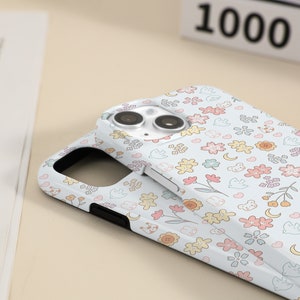 May include: A white phone case with a cute floral pattern featuring small flowers, hearts, moons, and birds. The case has a black bumper and a clear camera lens.
