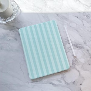 Puede incluir: Una funda para tablet azul claro con rayas blancas. La funda está sobre una superficie de mármol blanco.