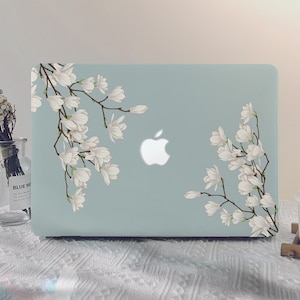 以下が含まれることがあります： 白い花柄のライトブルーのラップトップケース。ケースには、緑色の茎と葉を持つ白いマグノリアの花の枝が描かれています。アップルのロゴがケースに表示されています。