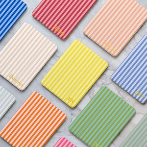 May include: A collection of colorful, striped rectangular cases. The cases are in various colors, including blue, pink, yellow, and orange. Some cases have gold lettering with names or initials. The cases are arranged on a marble surface.