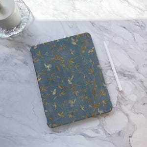 Puede incluir: Una funda azul para tableta con un diseño de ramas doradas y pequeños pájaros blancos. Un lápiz óptico blanco descansa junto a la funda sobre una superficie de mármol. La funda es rectangular.